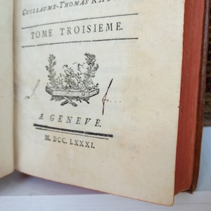 May include: Open book with text in French, including "TOME TROISIEME" and "A GENEVE." The book has a red spine and aged, cream-colored pages. A decorative emblem is printed on the page. The book appears to be antique.