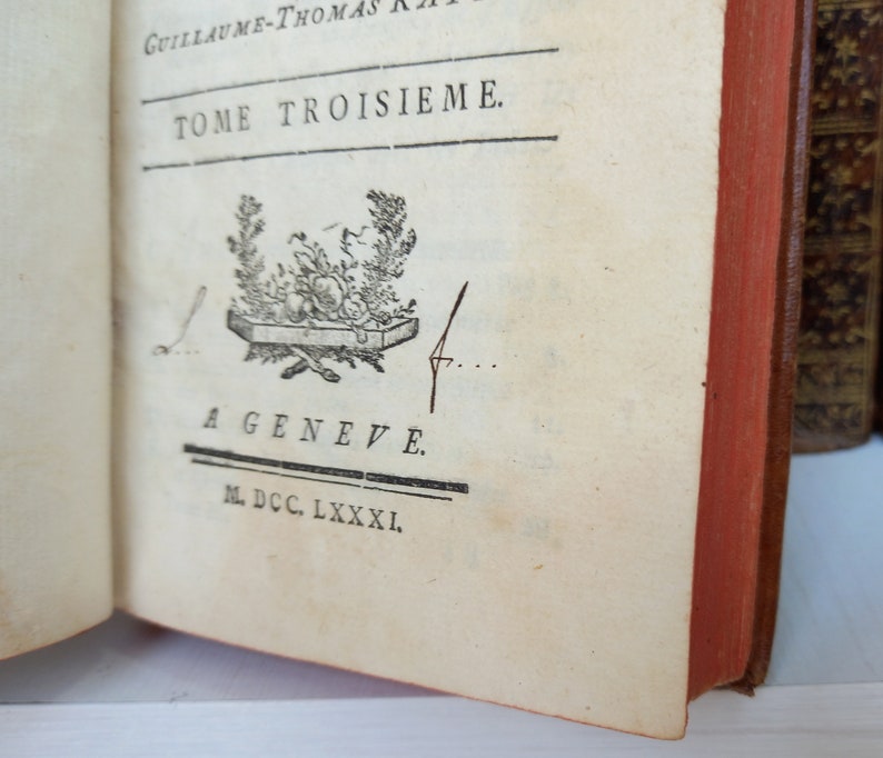 May include: Open book with text in French, including "TOME TROISIEME" and "A GENEVE." The book has a red spine and aged, cream-colored pages. A decorative emblem is printed on the page. The book appears to be antique.