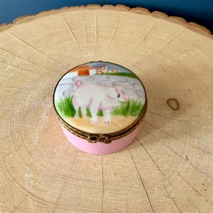 May include: A small, round, pink trinket box with a painted lid. The lid features a pink pig standing in a grassy field with a farm scene in the background. The box has a gold-colored metal trim and clasp.