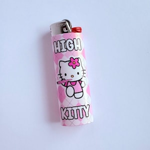 Puede incluir: Encendedor rosa y blanco con un patr&oacute;n de diamantes. El encendedor presenta una imagen de dibujos animados de Hello Kitty con una flor rosa y las palabras "HIGH KITTY" en negrita.