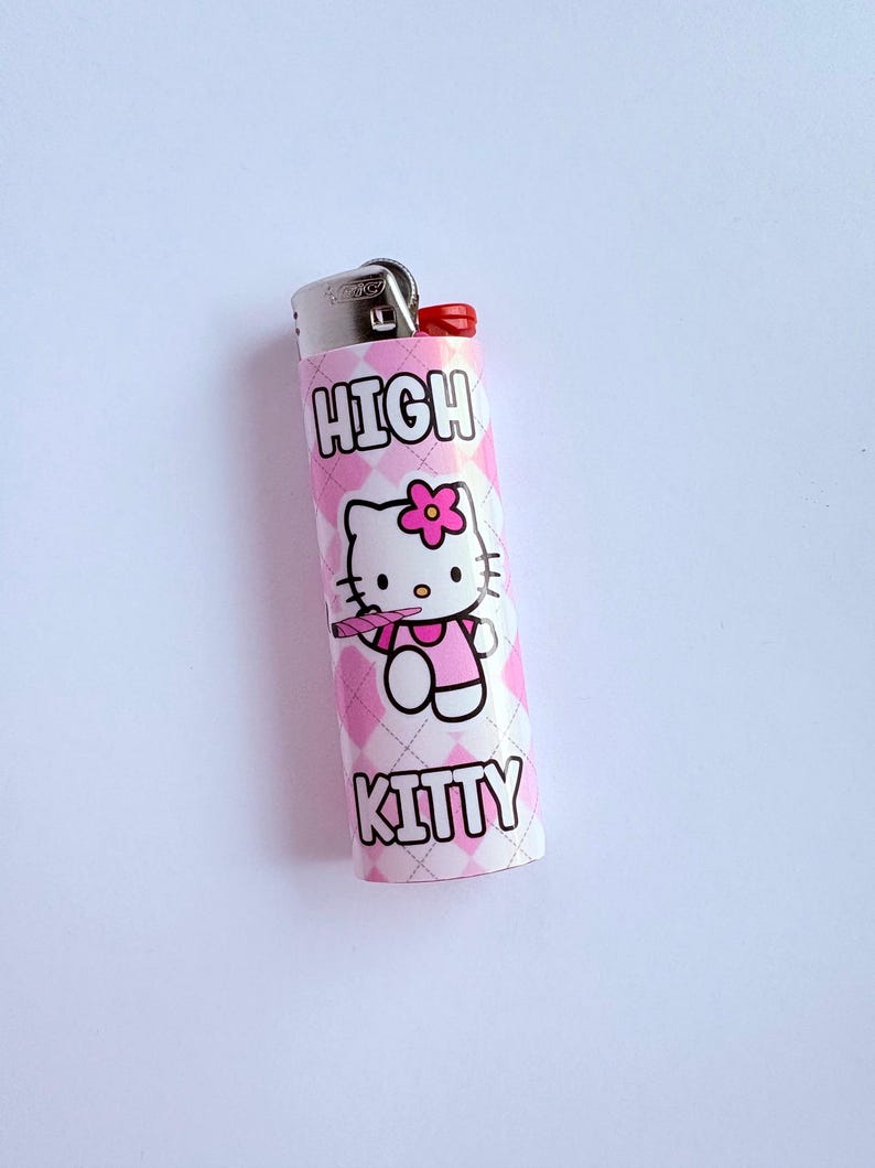 Puede incluir: Encendedor rosa y blanco con un patr&oacute;n de diamantes. El encendedor presenta una imagen de dibujos animados de Hello Kitty con una flor rosa y las palabras "HIGH KITTY" en negrita.