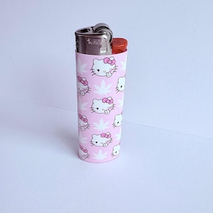 Puede incluir: Una funda de encendedor rosa con un patr&oacute;n repetido de caras blancas de Hello Kitty con lazos rosas y hojas de cannabis blancas. El encendedor es plateado con un bot&oacute;n rojo.