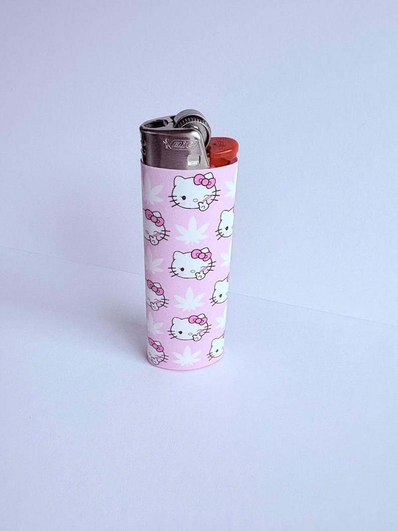Puede incluir: Una funda de encendedor rosa con un patr&oacute;n repetido de caras blancas de Hello Kitty con lazos rosas y hojas de cannabis blancas. El encendedor es plateado con un bot&oacute;n rojo.
