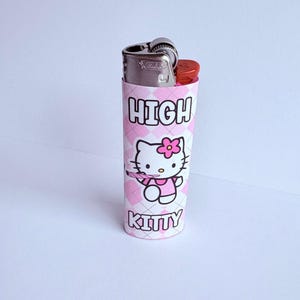 Puede incluir: Una funda para encendedor con estampado de diamantes rosas y blancos con las palabras "HIGH KITTY" impresas. La funda presenta una imagen de dibujos animados de Hello Kitty sosteniendo una flor. El encendedor es plateado y rojo.
