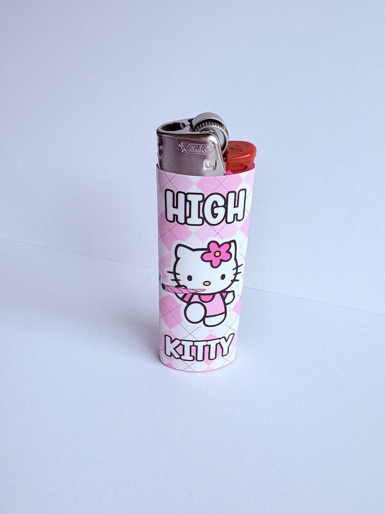 Puede incluir: Una funda para encendedor con estampado de diamantes rosas y blancos con las palabras "HIGH KITTY" impresas. La funda presenta una imagen de dibujos animados de Hello Kitty sosteniendo una flor. El encendedor es plateado y rojo.