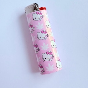 Puede incluir: Encendedor rosa con un patr&oacute;n repetido de caras de Hello Kitty y hojas de cannabis. El encendedor tiene una parte superior de metal plateado y un bot&oacute;n de encendido rojo.