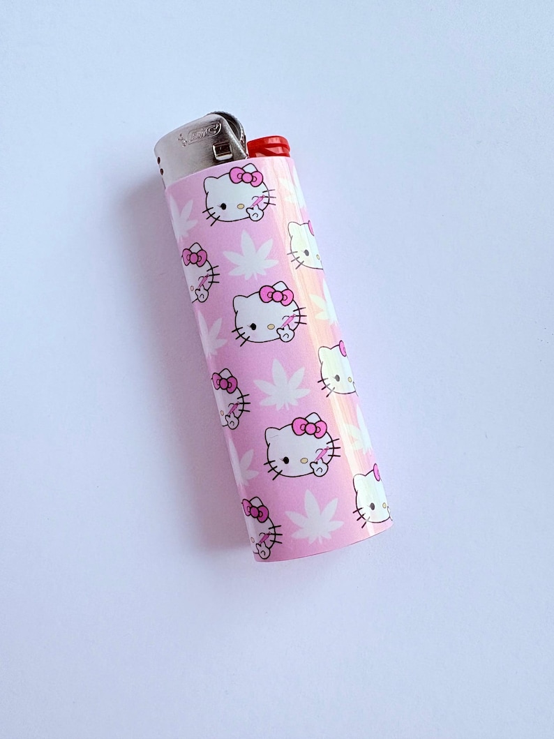 Puede incluir: Encendedor rosa con un patr&oacute;n repetido de caras de Hello Kitty y hojas de cannabis. El encendedor tiene una parte superior de metal plateado y un bot&oacute;n de encendido rojo.