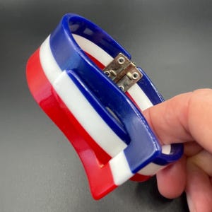 Peut inclure: Un bracelet articulé rouge, blanc et bleu. Le bracelet a une forme incurvée avec un fermoir en métal. Les couleurs sont disposées en rayures, avec le rouge en bas, le blanc au milieu et le bleu en haut.