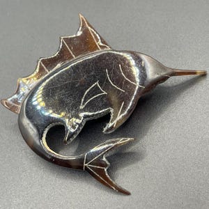 Puede incluir: Broche vintage con forma de pez y acabado oscuro iridiscente. El broche presenta un diseño detallado con aleta dorsal, cola y hocico puntiagudo. El pez mide aproximadamente 7.6 cm de largo y está hecho de un material brillante, posiblemente metálico.