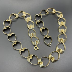 May include: A gold-toned necklace featuring a series of heart-shaped links. The necklace has a delicate design with a clasp closure. The hearts are evenly spaced and create a romantic and elegant look. The necklace is displayed on a dark surface.
