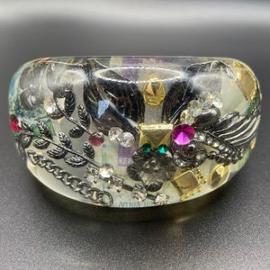 Pode incluir: Um bracelete de resina transparente com uma borda dourada e incrustações de enfeites pretos, dourados e verdes. O bracelete apresenta uma corrente, folhas e pedrarias.