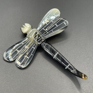 May include: A detailed dragonfly sculpture with translucent wings and a segmented body. The wings are a light gray with a grid-like pattern, while the body transitions from dark gray to a yellowish-brown tip. The sculpture is set against a dark gray background.