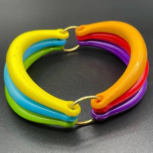 Puede incluir: Un brazalete de plástico multicolor con un anillo de metal dorado. El brazalete está hecho de cinco bandas de diferentes colores: amarillo, verde, azul, rojo y morado.