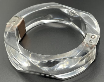 Puede incluir: Un brazalete de plástico transparente con detalles en metal plateado. El brazalete está abierto y tiene un diseño facetado.