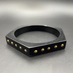 May include: A black hexagonal bangle bracelet featuring a series of small, round, metallic gold-toned studs embedded along the outer edge. The bracelet has a smooth, matte finish and a geometric shape with six distinct sides and corners.