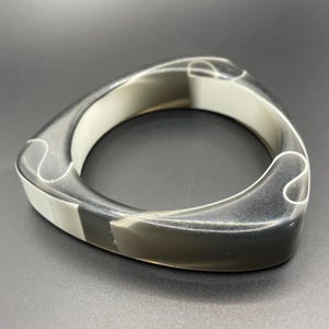 May include: A geometric bangle bracelet in a triangular shape. The bracelet is made of translucent gray and white material. The design features curved edges and a smooth finish, suitable for everyday wear.