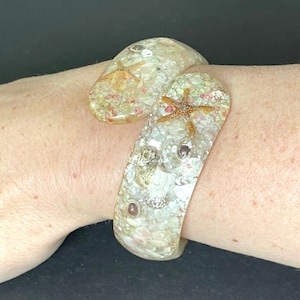 Puede incluir: Un brazalete blanco de resina con conchas marinas, estrellas de mar y purpurina iridiscente incrustados.