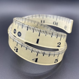 May include: A coiled, cream-coloured measuring tape with black markings. The tape is marked in inches, with numbers from 0 to 12 visible. The tape is made of a flexible material and is designed for measuring.