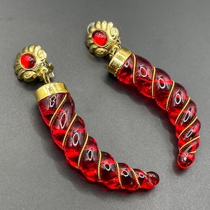 May include: A pair of drop earrings featuring gold-tone metal tops with a circular red cabochon stone. Each earring has a long, curved, translucent red glass horn shape wrapped in a thin gold-tone wire spiral, creating a distinct twisted design.