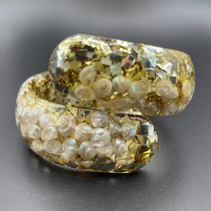Puede incluir: Una pulsera de resina transparente con un diseño en espiral. La pulsera está llena de pequeñas conchas blancas y purpurina dorada. Una joya única.