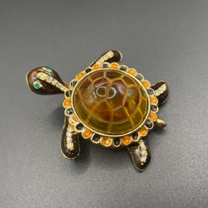May include: A decorative turtle brooch with a brown body, amber shell, and gold-colored accents. The brooch features clear and green gemstones, adding sparkle to the design. The turtle's shell has a honeycomb pattern.