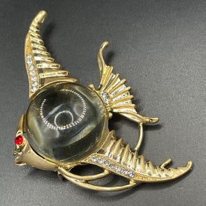 May include: Gold-coloured fish brooch with a large, clear, spherical centre. The brooch features a red jewel eye and is embellished with clear rhinestones. The fish has detailed fins and a curved tail, creating a unique accessory.