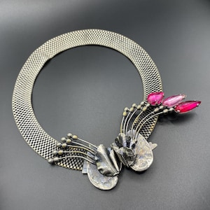 Peut inclure: Un collier argenté avec un bracelet en maille et des détails floraux. Le collier présente des pierres roses en forme de goutte et des feuilles métalliques. Le design comprend de petites perles rondes argentées et des détails en métal.