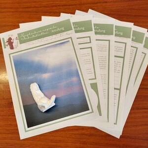 May include: A collection of white papers with green detailing and text, displayed on a wooden surface. One paper shows a white sculpture against a blue and purple backdrop. The papers feature German text and a small illustration.