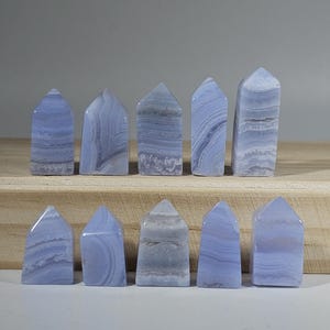 May include: A collection of ten blue lace agate crystal towers. The crystals are various sizes and shapes, with a banded pattern of light and dark blue. They are arranged on a wooden surface against a neutral background.