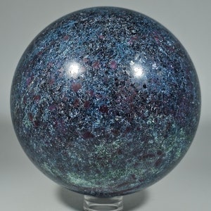 May include: A spherical decorative object with a speckled pattern in shades of blue, green, and purple. The sphere is resting on a clear stand. The surface has a textured appearance, with small, irregular spots of color.