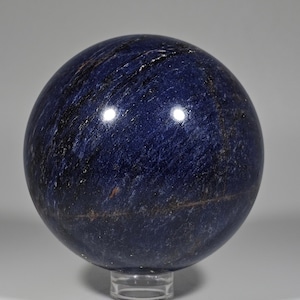 May include: A polished, spherical gemstone in deep blue with streaks of black and brown. The sphere is displayed on a clear, circular stand. The surface reflects light, highlighting the stone's texture and color variations. A decorative item.