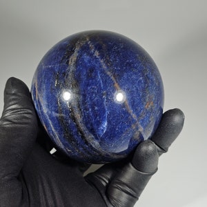 May include: A polished, spherical sodalite gemstone. The sphere is a deep blue color with streaks of white, black, and gold. The gemstone is held in a black gloved hand against a plain white background.