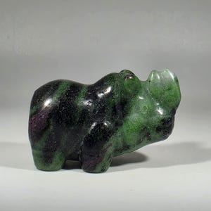May include: A carved stone figurine of a rhinoceros. The sculpture is primarily dark green with streaks of black and patches of purple. The rhino is posed in a standing position.