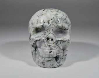 May include: A carved skull figurine in white and gray. The skull has detailed features, including eye sockets, a nose cavity, and a full set of teeth. The surface has a marbled appearance with dark gray veining.