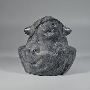 May include: A gray stone sculpture of a creature with a smiling face, small ears, and paws. The sculpture is seated in a bowl-like form. The stone has a textured appearance with visible veining.