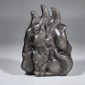 May include: A dark gray sculpture of a squirrel with a textured surface. The squirrel is sitting with its tail curled up, and the sculpture has a flame-like shape behind it. The sculpture is made of a shiny, reflective material.