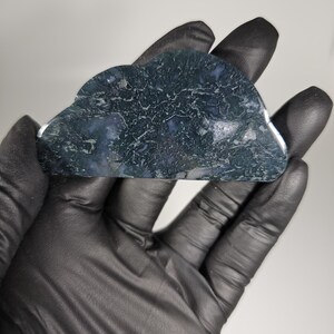 May include: A cloud-shaped stone with a dark green and blue pattern. The stone is held in a gloved hand against a white background. The stone's surface has a textured appearance, with intricate veining and color variations.