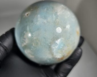 May include: A light blue sphere with a marbled appearance, held in a gloved hand. The sphere has a mix of blue, white, and tan colors, with a smooth, polished surface. The object is a decorative item, possibly a crystal or stone.