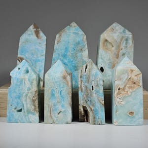 May include: A collection of seven blue aragonite crystal towers with brown and white veining. The crystals are displayed on a wooden block against a gray background. Each tower has a pointed top.