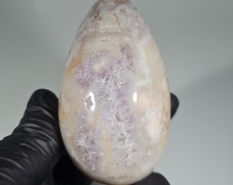 Op de afbeelding: Een gepolijste, eivormige steen met een mix van kleuren, waaronder lavendel, crème en hints van oranje. De steen heeft een glad, glanzend oppervlak en een kristalachtig interieur. Het object wordt vastgehouden door een gehandschoende hand.