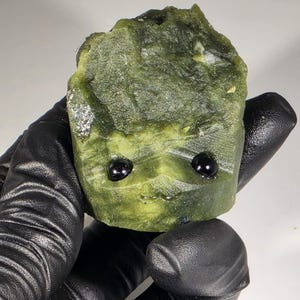 May include: A green stone carving of a character with two black eyes and a small smile. The stone has a rough, textured surface and is held by a gloved hand. The carving is a unique decorative item.