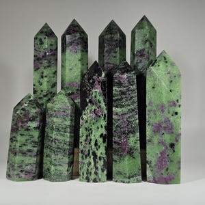 May include: A collection of polished ruby zoisite crystal towers. The crystals display a mix of green, black, and pink hues. The towers vary in height and shape, with pointed tops. The natural stone formations are arranged against a neutral background.