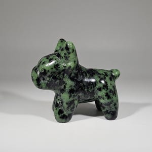May include: A small, green and black speckled stone figurine of a dog. The dog is standing on four legs with its ears perked up. The figurine is smooth and polished, with a glossy finish.