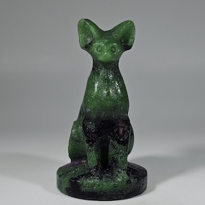 May include: A green and black cat figurine. The cat is sitting upright with its ears perked up. The figurine has a smooth, glossy finish and is set on a round base. The cat's body transitions from green to black.