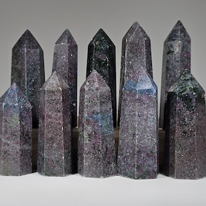 May include: A collection of ten dark gray, purple, and green gemstone points, also known as kyanite, arranged in a row on a wooden surface.