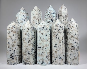 May include: A collection of ten K2 stone obelisks. The obelisks are white with black and gray speckles and a light blue hue. Each obelisk has a pointed top and a rectangular base. The stones are arranged in a group, with some in the front and some in the back.