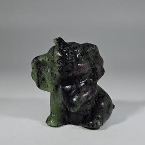 May include: A small, carved stone elephant figurine in shades of green and purple. The elephant is depicted in a seated position with its trunk down. The stone has a speckled appearance.