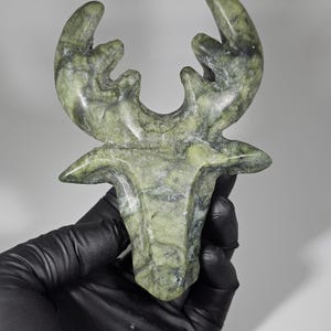 May include: A green and gray stone sculpture of a moose head with antlers. The sculpture is held by a gloved hand against a white background. The stone has a marbled appearance with variations in color.