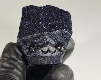 May include: A dark blue, glittery stone with a carved face. The stone is held by a gloved hand. The stone has a rough, textured surface and a sparkling appearance, with small flecks of silver and blue. The carved face has small, round eyes and a smiling mouth.
