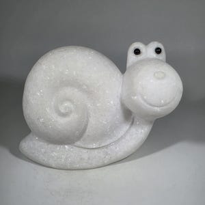May include: A white, whimsical snail sculpture with a spiraled shell and a smiling face. The snail has black eyes and a smooth, rounded body. The sculpture is made of a material that appears to have a crystalline texture.